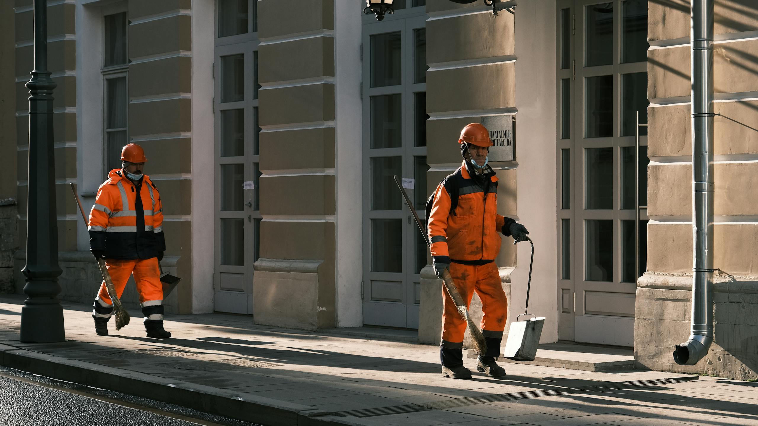 Two Men in Orange Workwear Walking on Sidewalk Holding Broomsticks and Dustpans